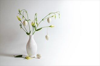 Delicate flower bunch in a white vase decorated with hanging Easter eggs on a light gray