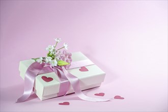 Gift box with flower bouquet, ribbon and paper heart shapes on a light pink background, love