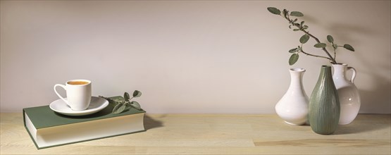 Coffee cup on a gray green book and a group of vases with a sage leaf branch on a wooden table