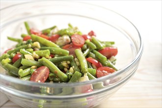 Salad of green beans, tomatoes, pine nuts and feta cheese in a glass bowl on a light wooden table,