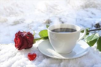 Hot coffee in a white cup served outdoors in the snow, decorated with a red rose and a little heart