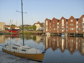 A harbour with a sailing boat and red brick facades in the calm water in the evening light,