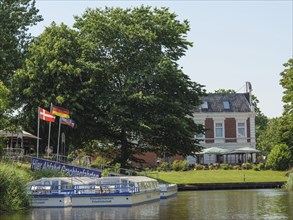 A tourist boat on the canal, surrounded by green trees and a historic villa with flags in the