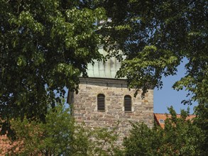 A stone church tower, surrounded by dense tree foliage, under a clear summer's day sky,