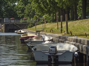 Boats along a canal, moored to wooden piles, shore with trees under a summer sky, friedrichstadt,