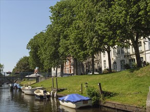 Boats along a canal in a green setting with lawn and trees, white houses in the background under a