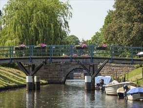 Wooden bridge over the canal, lined with flowers, boats on the water and green trees in the