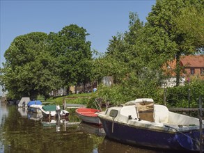 Boats moored along a riverbank, surrounded by lush greenery and trees on a sunny day,