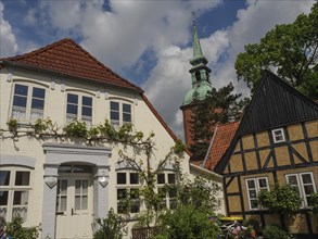 A historic street with half-timbered houses, flowers and a church tower under a cloudy sky with