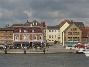 Historic buildings and tourists along a harbour shore under a cloudy sky, Kappeln,