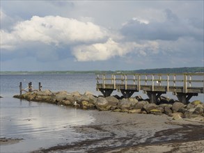 Wooden footbridge leading into the sea, pile of stones on the coast, clouds in the sky,