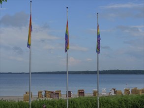 Beach with three rainbow flags, sea in the background, some clouds in the sky and beach chairs,