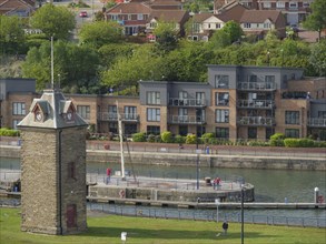 Stone tower on the riverside with modern residential buildings in the background, people walking on