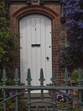 White door in a brick façade, surrounded by flowers and a green metal fence, charming and historic,