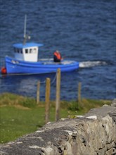 A blurred fishing boat and fence are seen behind a stone wall over blue water, Lerwick, Shetlands,