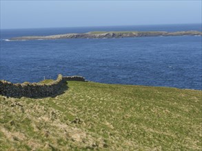 Green meadow with stone wall on the shore that merges into the wide blue sea and visible islands,