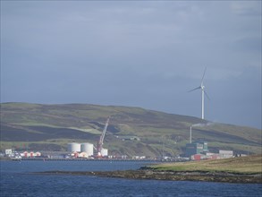 Coastal landscape with green hills, wind turbine and industrial buildings on the horizon, Lerwick,