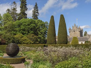 Green garden with hedges, plants and a sculpture, in the background a stone castle under a blue