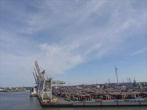 Industrial harbour with stacks of containers and several cranes under a partly cloudy sky, Hamburg,