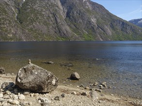 A large boulder lies on a rocky beach with a tranquil fjord and mountains beyond, Eidfjörd, Norway,