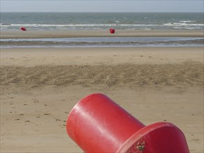 Close-up of a red buoy in the sandy beach area, with several buoys in the background and calm sea,