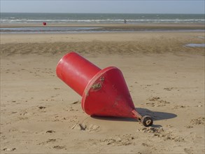 A large red buoy lies on the sand on the beach with the sea in the background, de haan, belgium