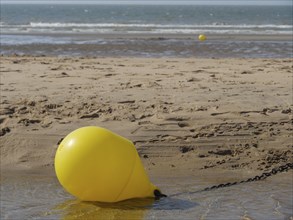 A large yellow buoy lies in the sand on the beach with calm water in the background, de haan,