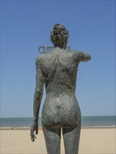 Bronze statue of a naked pregnant woman on the beach, overlooking the sea under a clear blue sky,