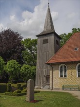 Historic wooden tower next to a sand-coloured church, surrounded by lush greenery, Arnis,