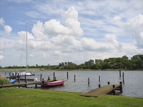 A calm harbour picture with boats at the jetty and a cloudy sky above the trees in the background,