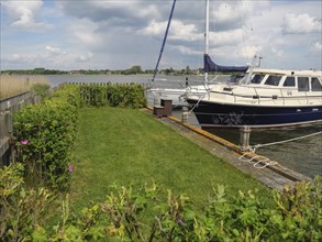 A sailing boat moored to a wooden jetty, surrounded by bushes and a cloudy sky, Arnis,