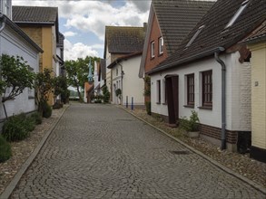 A quiet cobbled street with stylish houses and trees under a cloudy sky, Maasholm,