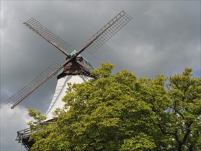 A large windmill rises into the sky, surrounded by dense green trees and a cloudy sky, Kappeln,