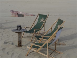 Two green deckchairs and a small table on a sandy beach, small boat in the background, Juist, East