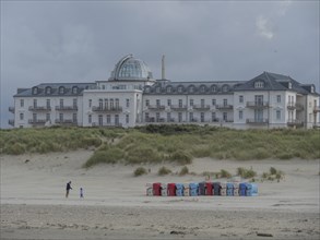 Large hotel with dome in the background, in front of it sandy beach with some people, cloudy sky,