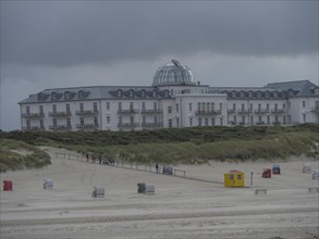 Large hotel building with a dome in the foreground, people walking on a sandy beach, juist, east