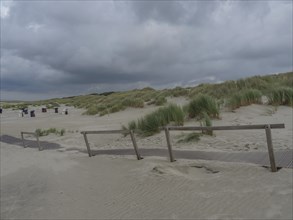 Beach shot with wooden path, dunes, sand and cloudy sky, scattered beach chairs in the background,