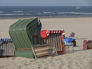 Several colourful beach chairs stand on the sandy beach in front of the sea, a sailing boat can be