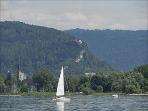 A sailing boat on a calm lake, surrounded by green hills and a church, Lindau, Lake Constance,