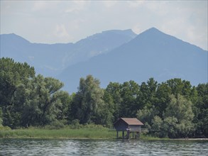 Small wooden hut on the shore of a lake, surrounded by trees with a mountain backdrop, Lindau, Lake