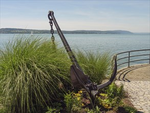 Large anchor on the lakeshore, surrounded by grass and flowers with a view of the calm lake,