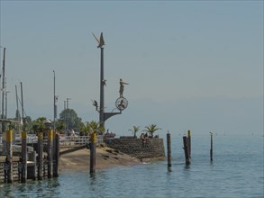 Sculptures on the lakeshore next to a jetty, in the background the calm water and the blue sky,