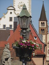 A historic street lamp with flowers in front of old buildings and a church tower under a blue sky,