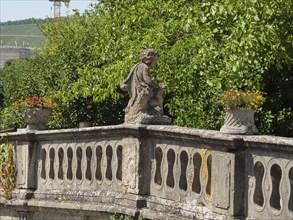 Stone statue on an old balcony railing, surrounded by green trees and plants, a touch of history