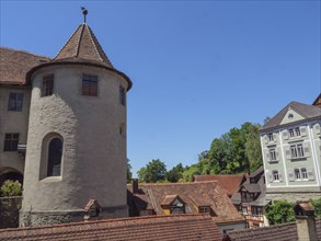 A castle with a round tower and surrounding roofs of historic buildings under a clear blue sky,