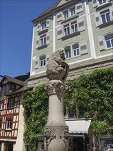 Statue on a column in front of historic buildings with green facades and ivy in a sunny old town,