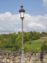 Lonely historic street lamp in front of a stone wall and a green landscape with trees and blue sky