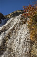 Water flows down a rocky slope, accompanied by colourful autumn leaves under a bright blue sky,