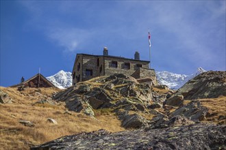 A mountain hut stands on a rocky slope with a distant snow-capped mountain range in the background,