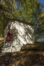 A white chapel stands in the forest under sunlight, surrounded by trees and shade, Saas Fee,